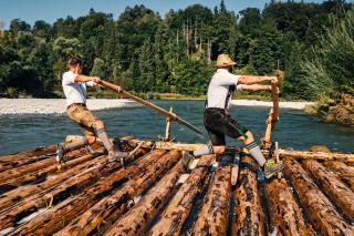 Auf dem Bild sieht man zwei Männer, die gerade auf einem Holzfloß im Wasser mit einem Holzruder rudern. Beide tragen eine Tracht. 